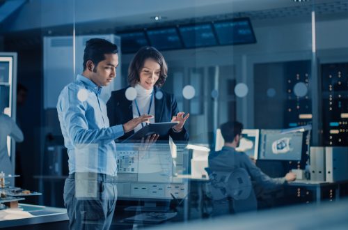 In Technology Research Facility: Female Project Manager Talks Wi In Technology Research Facility: Female Project Manager Talks With Chief Engineer, they Consult Tablet Computer. Team of Industrial Engineers, Developers Work on Engine Design Using Computers